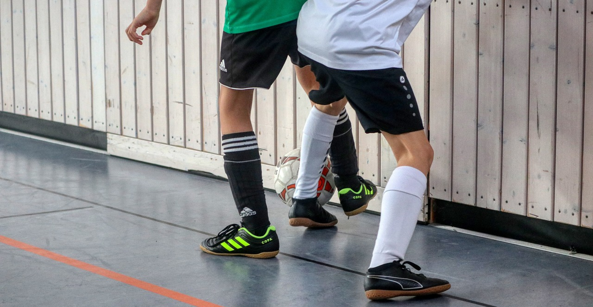 Zwei Hallenfußballspieler im Zweikampf nahe der Bande, mit schwarz-weißen Trikots und Sportschuhen auf dem Hallenboden.